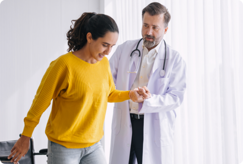 Ein Arzt hält die Hand einer Frau, die aus einem Rollstuhl aufsteht. 