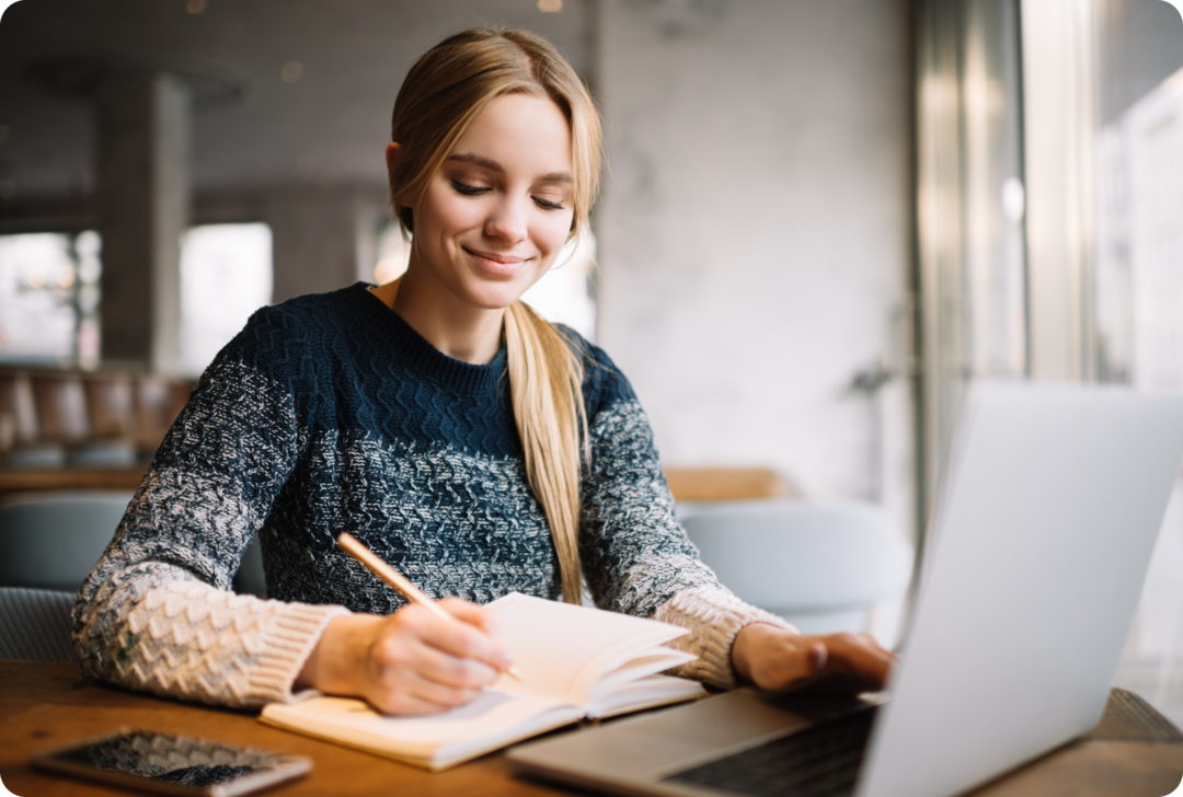 Eine Frau tippt mit der linken Hand auf der Tastatur eines Notebooks, mit der rechten Hand schreibt sie in ein Notizbuch.