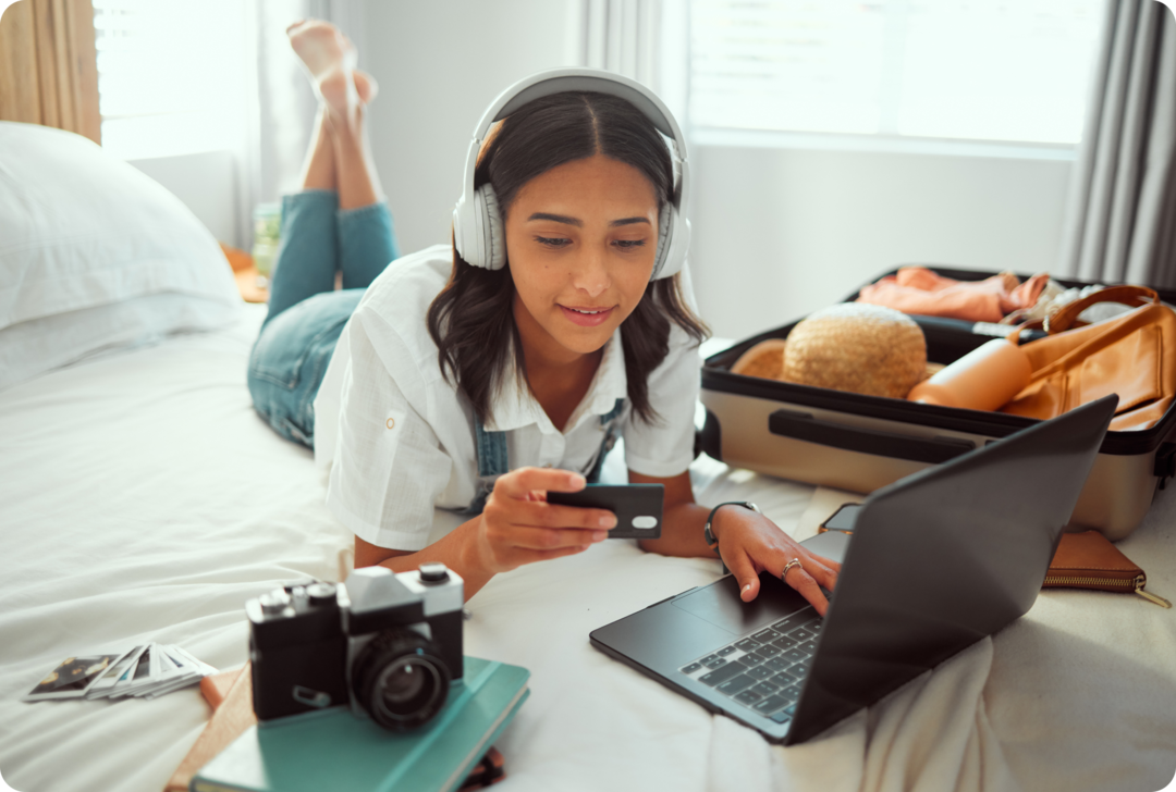 Urlaubsvorbereitung bei MS Eine junge Frau liegt auf einem Bett. Neben ihr ein geöffneter Koffer, eine Kamera und ein Laptop.