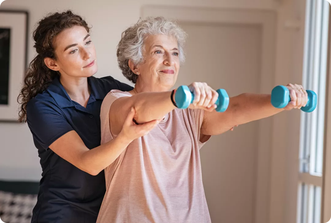 Eine ältere Frau hält zwei Hanteln vor sich. Eine jüngere Frau steht hinter ihr und unterstützt sie.