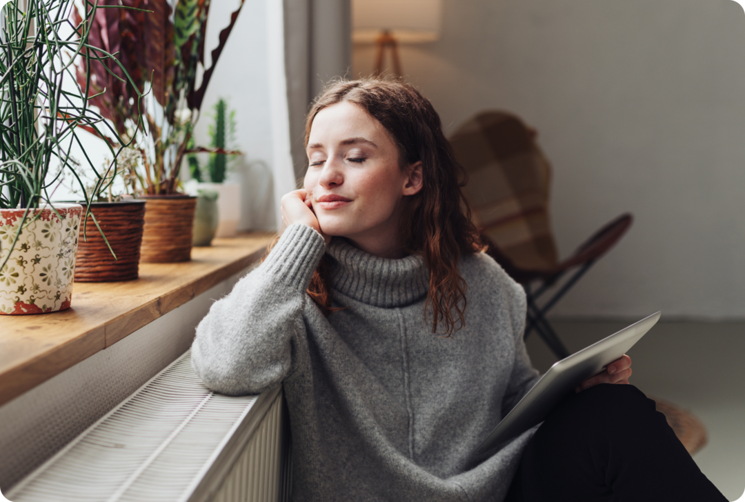 Eine Frau sitzt mit geschlossenen Augen vor einem Fenster und lehnt sich an einen Heizkörper.