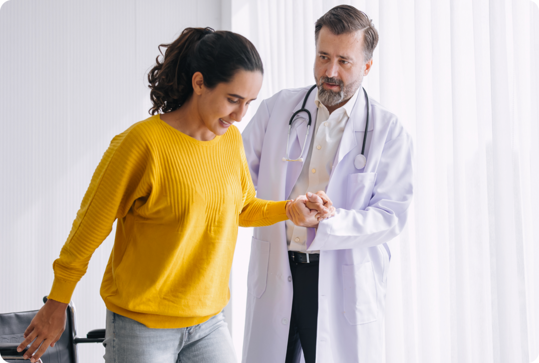 Ein Arzt hält die Hand einer Frau, die aus einem Rollstuhl aufsteht. 