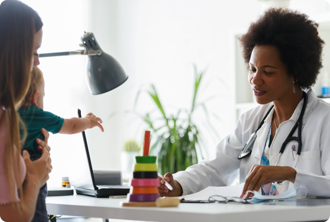 Eine Frau mit weißem Kittel und Stethoskop um den Hals schaut auf ein Dokument. Eine Frau mit einem Klainkind auf dem Arm sitzt ihr gegenüber.