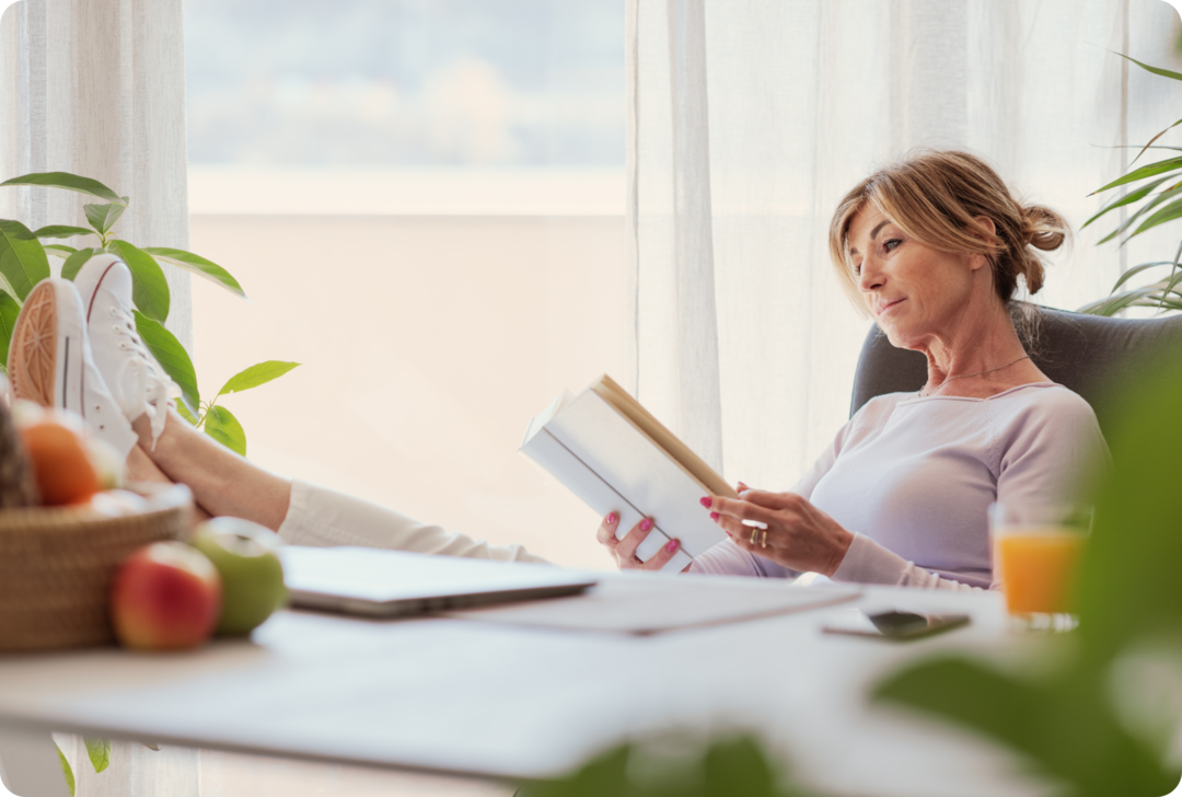 Eine Frau sitzt mit hochgelegten Beinen an einem Schreibtisch und liest in einem Buch.