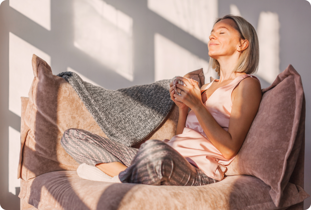 MS: Alltagshilfen Eine Frau sitzt mit überschlagenen Beinen auf einem Sofa. Sie hält mit beiden Händen eine Tasse und hat die Augen geschlossen. Sonnenstrahlen scheinen ihr ins Gesicht.