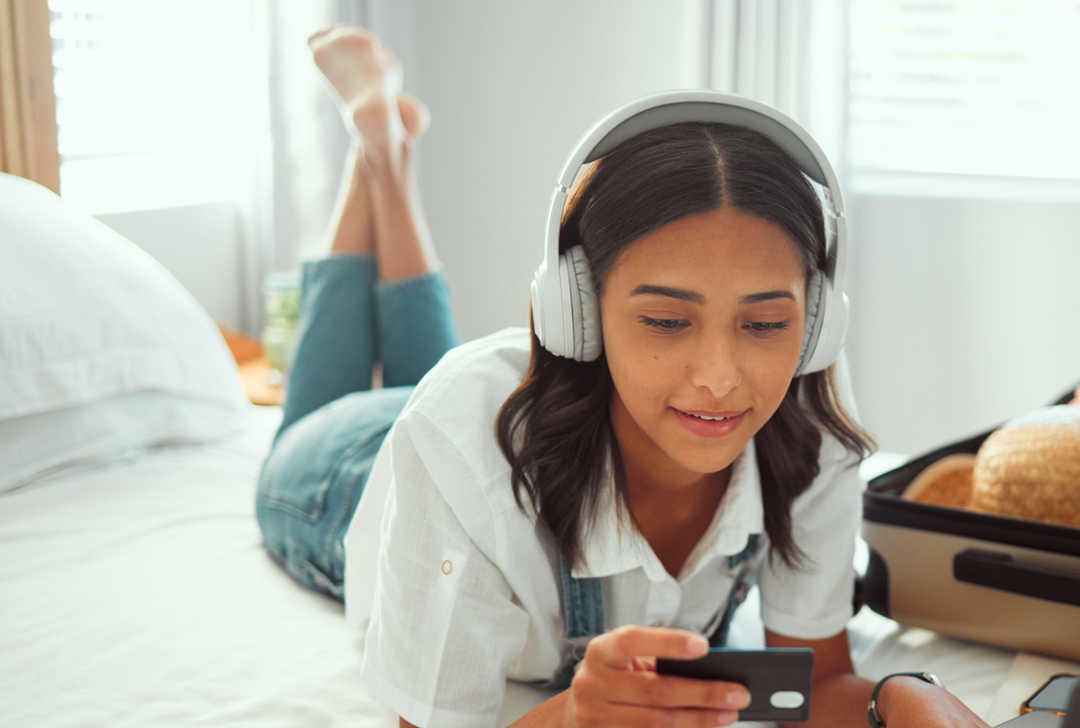 Eine junge Frau mit Kopfhörer auf den Ohren liegt auf einem Bett und schaut auf ihr Smartphone.